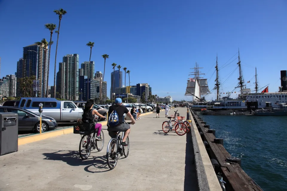 People biking along beach path with moored ship in San Diego, California.
